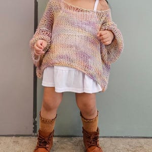 May include: A young child wearing a colorful, open-weave sweater in shades of yellow, pink, and purple. The sweater is paired with a white dress, brown socks, and brown leather boots. The outfit is suitable for a child.