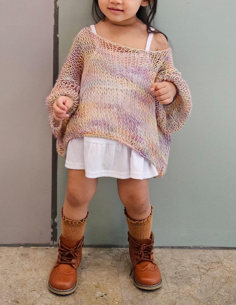 May include: A young child wearing a colorful, open-weave sweater in shades of yellow, pink, and purple. The sweater is paired with a white dress, brown socks, and brown leather boots. The outfit is suitable for a child.