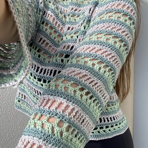 May include: A pastel-coloured crochet sweater with long, flared sleeves. The sweater features a mix of light green, blue, and white yarn, creating a striped and open-weave design. The neckline is a simple scoop, and the overall style is bohemian.