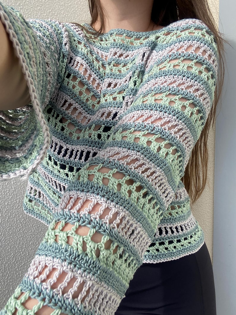 May include: A pastel-coloured crochet sweater with long, flared sleeves. The sweater features a mix of light green, blue, and white yarn, creating a striped and open-weave design. The neckline is a simple scoop, and the overall style is bohemian.