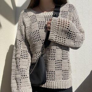 Pu&ograve; includere: Un maglione beige all'uncinetto con un motivo a trama aperta e ampia. Il maglione ha maniche lunghe e scollo tondo. &Egrave; visibile una borsa a tracolla nera con catena argentata.