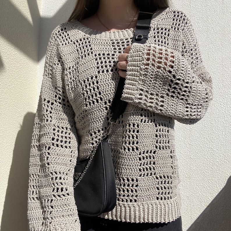 Pu&ograve; includere: Un maglione beige all'uncinetto con un motivo a trama aperta e ampia. Il maglione ha maniche lunghe e scollo tondo. &Egrave; visibile una borsa a tracolla nera con catena argentata.