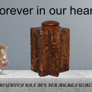 May include: A wooden urn box with a star-shaped base and a lid. The urn is made of rosewood and has a natural wood finish. The text "Forever in our hearts" is written above the urn. The text "ROSEWOOD URN BOX FOR HUMAN ASHES" is written below the urn.