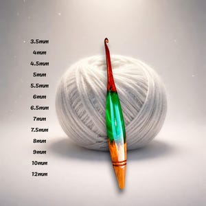 Op de afbeelding: Een haaknaald met een taps toelopende punt, voorzien van een rood en groen hars handvat dat overgaat in een natuurlijke houten basis met twee horizontale groeven. Een lijst met maten van 3,5 mm tot 12 mm staat links in de afbeelding.