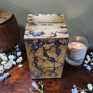 May include: Wooden urn with a natural wood grain finish and blue lightning-like patterns. The lid is engraved with the name "Donald Hausner" and the years "1943-2026". A lit candle in a glass jar sits nearby, with a floral arrangement.