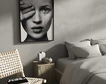 May include: A black and white framed print of a woman's face with her hand covering one eye. A woven chair with a book and a small wooden side table with a vase and decorative pumpkin are in the foreground. A bed with white bedding is in the background.