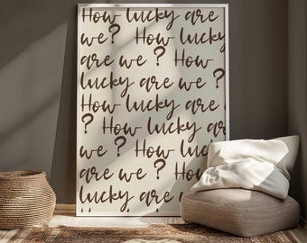 May include: A framed print with the text "How lucky are we?" repeated in a handwritten style. The print is in a white frame and leans against a wall. A woven basket, a patterned rug, and a beige pouf with pillows are in the foreground.