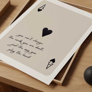 Op de afbeelding: Een beige kaart met een zwarte schoppen aas en de quote "Je kunt de kaarten die je krijgt niet veranderen, alleen de manier waarop je de hand speelt".