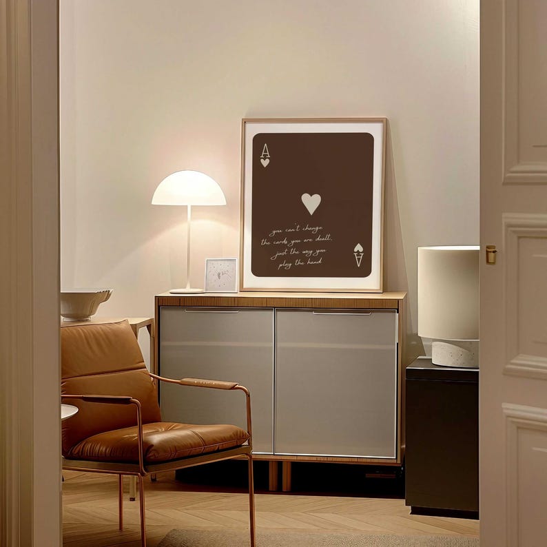 May include: A framed Ace of Hearts playing card art print with the quote "you can't change the cards you are dealt, just the way you play the hand." The print is brown with white text and a white border. A modern interior design theme.