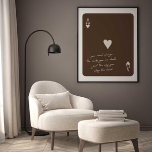 May include: A framed Ace of Hearts playing card art print with the text "you can't change the cards you are dealt, just the way you play the hand." The print hangs above a beige armchair and ottoman.