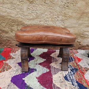 Könnte beinhalten: Ein brauner Lederhocker mit geschnitztem Holzsockel. Der Hocker hat eine abgerundete, gepolsterte Oberseite mit einem dekorativen Steppmuster. Die Holzbeine sind aufwendig geschnitzt. Der Hocker steht auf einem bunten Teppich.