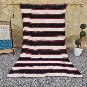 May include: A large, rectangular rug with horizontal stripes in black, white, and burgundy. The rug is displayed against a neutral-colored wall. The rug's design is simple and bold, suitable for various interior styles.