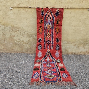 May include: A long, rectangular runner rug featuring a vibrant red base color with intricate geometric patterns in blue, yellow, orange, white, and black. The design includes two arched motifs and a fringed edge at the bottom end of the textile.