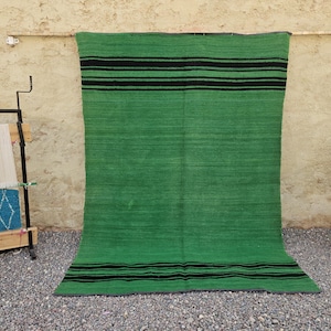 May include: A large rectangular rug featuring a vibrant green base colour with horizontal black stripes near the top and bottom edges. The rug has a flat-woven texture and is displayed hanging vertically against a textured wall surface.