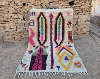 May include: A white Moroccan rug with a colorful geometric pattern. The rug features pink, yellow, green, blue, and purple accents. The rug has a fringe border.