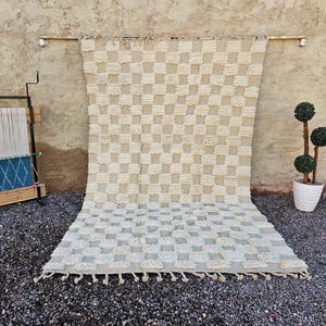 May include: A large, beige and white checkered rug with a textured, tufted design. The rug is displayed against a neutral-toned wall, with a small weaving loom and potted topiary nearby. The rug has a fringe along the bottom edge.