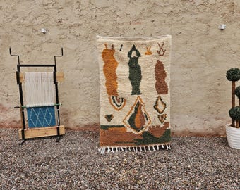 May include: A cream-colored, rectangular rug with geometric patterns in shades of brown, green, and orange. The rug has a fringe along the bottom edge. A weaving loom and a potted topiary are in the background.