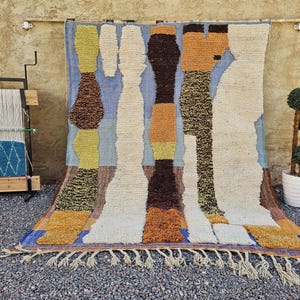May include: A large, colorful rug with an abstract design. The rug features vertical shapes in shades of brown, beige, yellow, and blue. It has a fringed edge and is displayed with a weaving loom and a potted topiary.