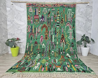 Könnte beinhalten: Ein lebendiger grüner Teppich mit einem farbenfrohen, abstrakten Design. Der Teppich weist ein sich wiederholendes Muster aus geometrischen Formen und Symbolen in verschiedenen Farben auf, darunter Rosa, Gelb und Blau. Der Teppich hat einen Fransenrand und wird vor einer neutralen Wand ausgestellt.