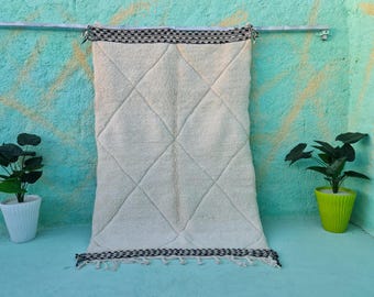 May include: A white Moroccan rug with a diamond pattern and black trim. The rug is hanging on a wall with a light blue background.