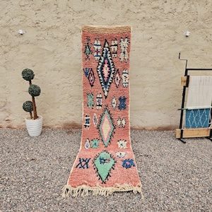 Op de afbeelding: Een lange, rechthoekige, handgeweven vloerkleed met een roze achtergrond en geometrische patronen in blauw, groen en wit. Het vloerkleed heeft een gefranjerde rand en wordt verticaal weergegeven. Een decoratieve plant en een weefgetouw staan op de achtergrond.