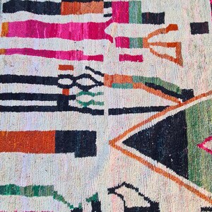 May include: A white rug with a geometric pattern in black, orange, pink, and green. The rug has a tribal design with a variety of shapes and lines.