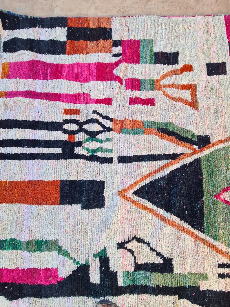 May include: A white rug with a geometric pattern in black, orange, pink, and green. The rug has a tribal design with a variety of shapes and lines.