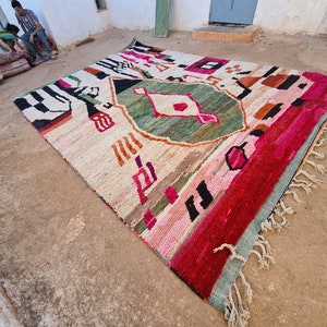 May include: A colorful, hand-woven rug with geometric patterns in shades of pink, red, green, and black. The rug has a fringe border and is laid out on a dirt floor.