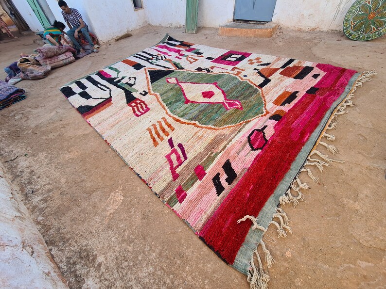 May include: A colorful, hand-woven rug with geometric patterns in shades of pink, red, green, and black. The rug has a fringe border and is laid out on a dirt floor.