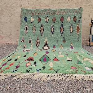 May include: A large, hand-woven, green rug with a geometric pattern. The rug features various shapes in colors like red, blue, and brown. It has a fringe along the bottom edge and is displayed outdoors.