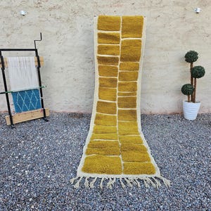 Könnte beinhalten: Rechteckiger Teppich in Senfgelb mit geometrischem Muster und cremefarbenem Fransen. Der Teppich wird auf einer Kiesoberfläche präsentiert. Ein Webstuhl und ein Topfbaum befinden sich im Hintergrund. Der Teppich ist ca. 200 cm lang.