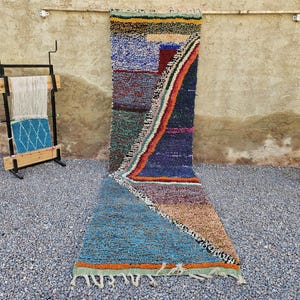 May include: A colorful, geometric Moroccan rug with a mix of blue, purple, orange, and green hues. The rug features a textured, woven design with a fringe along the bottom edge. A wooden loom and a potted plant are in the background.