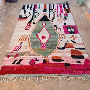 May include: A colorful Moroccan rug with a geometric pattern in shades of red, pink, green, black, and white. The rug has a diamond-shaped design in the center and a fringe border.