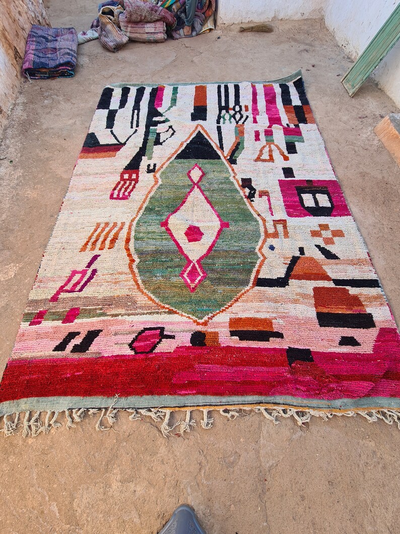 May include: A colorful Moroccan rug with a geometric pattern in shades of red, pink, green, black, and white. The rug has a diamond-shaped design in the center and a fringe border.