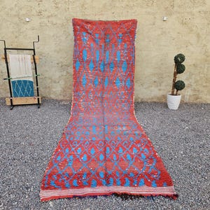 May include: A long, rectangular runner rug featuring a vibrant red base color with a repeating pattern of bright blue diamond shapes and abstract motifs. The rug has a textured surface and a horizontal striped border at the bottom edge.