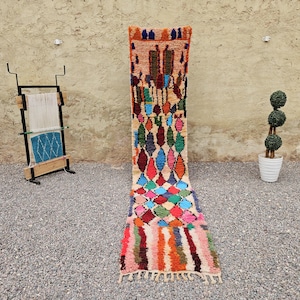 May include: A long, narrow runner rug featuring a cream-colored base with a vibrant, multi-colored diamond pattern in shades of red, blue, green, and orange. The rug ends in a series of vertical stripes and a fringe at the bottom edge.