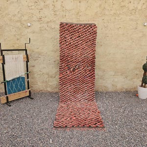Puede incluir: Una alfombra pasillera larga y rectangular con un pelo grueso y tupido en tonos rosa empolvado y marrón oscuro. La alfombra presenta una superficie densa y texturizada con un patrón repetitivo de pequeños mechones en relieve.