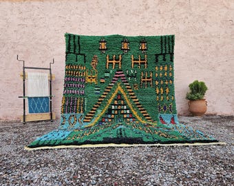 Könnte beinhalten: Ein lebendiger grüner Teppich mit geometrischen Mustern in Gelb, Schwarz und Blau. Der Teppich hat ein zentrales Dreiecksmuster und verschiedene rechteckige Formen. Eine kleine Pflanze in einem Terrakottatopf und ein Webstuhl befinden sich im Hintergrund.