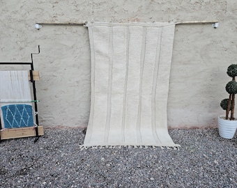 May include: A cream-colored, vertically striped rug with a fringed bottom hangs from a wooden rod. A weaving loom with white and blue yarn sits to the left. A topiary tree in a white pot is on the right.