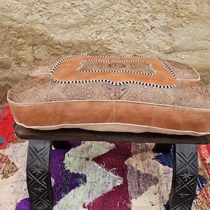 Peut inclure: Un pouf en cuir marron avec des motifs en relief complexes et une bordure blanche. Le pouf rectangulaire repose sur un tabouret en bois foncé. Le design présente des motifs géométriques et floraux, créant un élément décoratif et fonctionnel.
