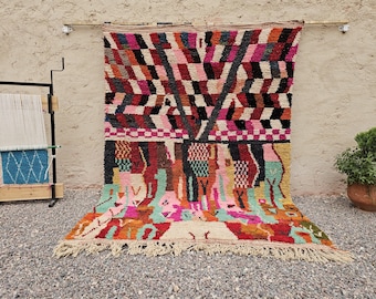 May include: A vibrant, hand-woven rug with a geometric pattern in shades of pink, black, red, and white. The rug features a fringed edge and a colorful, abstract design. A wooden loom and potted plant are in the background.