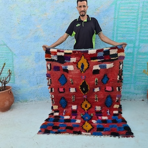 Pode incluir: Um tapete marroquino colorido com fundo vermelho e padrões geométricos em tons de azul, amarelo e branco. O tapete é tecido à mão e tem uma borda franjada.