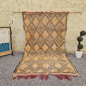 May include: A large rectangular rug featuring a golden-tan background with a repeating black diamond pattern. The rug has a dark red fringe along the bottom edge and a matching dark red border at the top, showcasing a geometric design throughout.