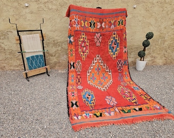 May include: A large rectangular rug featuring a vibrant orange background with a central diamond pattern in white, blue, and yellow. The rug is decorated with various geometric shapes and colorful motifs in pink, green, and black, finished with fringed edges.