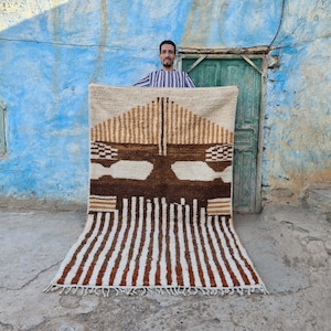 Könnte beinhalten: Ein Mann hält einen großen, rechteckigen Teppich mit einem geometrischen Muster in Braun-, Weiß- und Beigetönen hoch. Der Teppich hat einen Fransenrand und ist aus dickem, strukturiertem Garn gewebt.