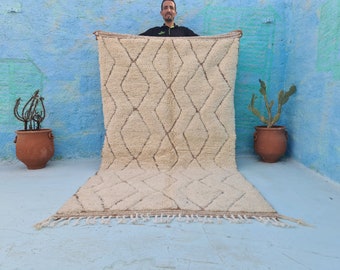 May include: A white and brown Moroccan rug with a diamond pattern. The rug has a fringe border and is being held up by a person.