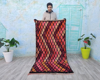 May include: A vibrant, hand-woven rug with a diamond pattern in shades of red, pink, orange, and black. The rug is displayed against a textured wall with a painted design. The rug's design and colors create a warm and inviting aesthetic.