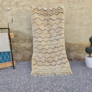 May include: A rectangular, cream-coloured wool rug featuring a repeating pattern of horizontal, dark brown zigzag lines. The rug is displayed vertically, showcasing its thick, textured pile and fringed edges at the bottom, with a consistent design.