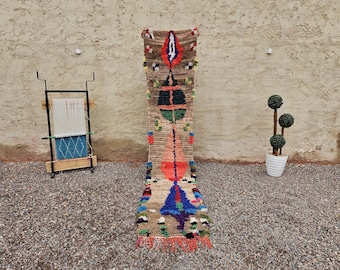 May include: A long, rectangular, hand-woven rug with a central design in shades of brown, red, blue, and green. The rug has a fringed edge and is displayed against a textured wall. A weaving loom and potted topiary are in the background.