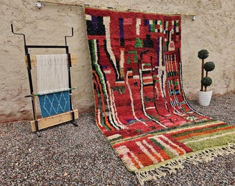 May include: A vibrant, hand-woven rug with a red base and a geometric design in various colors, including green, white, and black. The rug is displayed on a wall, with a weaving loom and a potted plant nearby. The rug has a fringed edge.